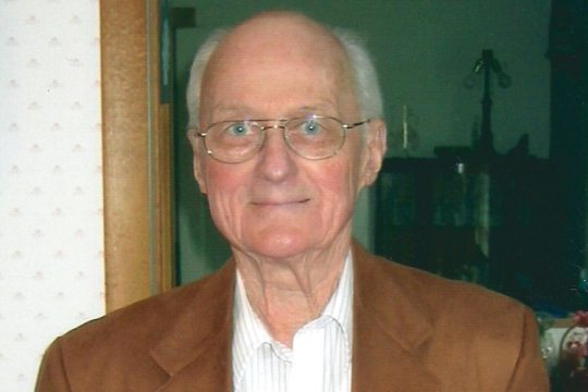  A color image. Headshot of an older man with thinning gray hair wearing glasses, an open-necked shirt, and a dark blazer. 