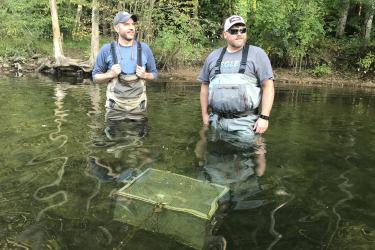 Two scientists stand in a stream, next to water quality testing equipment under the surface.