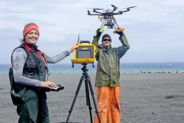 Photo of Katie Sweeney, holding a remote control console, and Brian Fadely, holding a drone, on a Bogoslof Island beach..