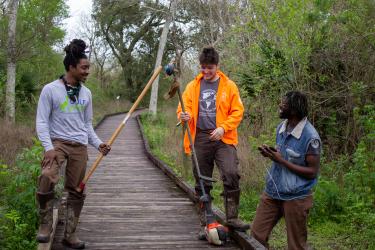 Three young people standing on a boardwalk in the woods with landscaping equipment.