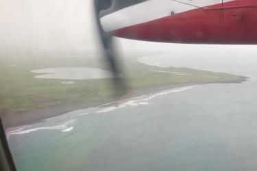 Aerial photo of island coastline with sandy beaches, green carpeted interior and small lakes from an aircraft window descending through a grey cloud ceiling.
