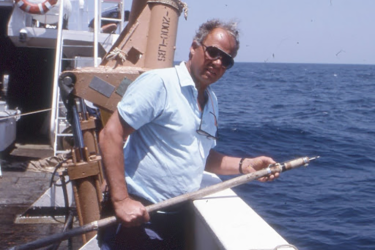 A scientist on the deck of a research ship at sea on a sunny day. He is holding a long pole with a sharp point at the end, used to insert tags into a shark’s fin. 