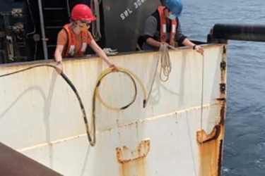 Two researchers on the deck of a research ship at sea wearing summer clothing, hard hats, and cloth masks to protect against COVID-19 transmission. They are easing an underwater sound recorder attached to a cable over the side and into the water. It is daylight and the weather is calm. 