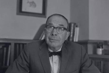 A black and whote photograph of a middle aged man at a desk with half-height bookcases filled with books in the background. The man wears glasses, a sport jacket with a light plaid shirt and dark, slender bow tie. 