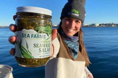 A smiling Atlantic Sea Farms employee stands on a dock while holding up a jar of 'Atlantic Sea Farms Fermented Seaweed Salad.'