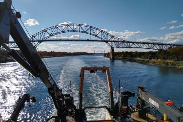  Color image taken during daylight from the upper deck of a research vessel, facing toward the rear of the vessel as it sails through a canal. Land is visible on each side of the canal, and the ship leaves a wake in the water. Two steel framed bridges are in the image. The ship has just passed under the bridge used by vehicles. The railroad bridge is in the distance. 