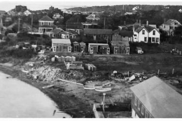 Three pigeon coops and sheds with screening are visible at the edge of a parking lot at center. View north shows private homes in the background looking toward Buzzards Bay. Great Harbor is at left. Materials and some small boats on the edge of the parking lot near the harbor at left. Photo: Courtesy Woods Hole Historical Museum Archives. 