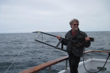Jay Barlow standing on a boat on the ocean, holding a tetrahedral metal frame with hydrophones attached. 