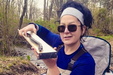 Valerie Ouellet wears a blue shirt and tan chest waders by a freshwater stream in a forest and holds a measuring board with an American eel on it. 