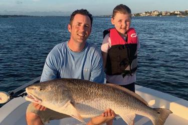A smiling father sits next to his young son near the bow of a recreational fishing boat, holding a large red drum in his hands.