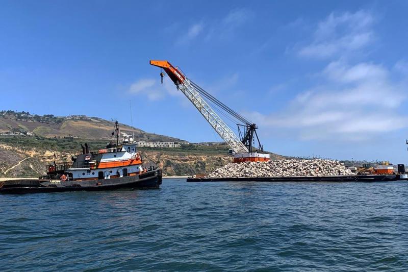 A tug boat approaching large barge with a pile of boulders off the California coast.