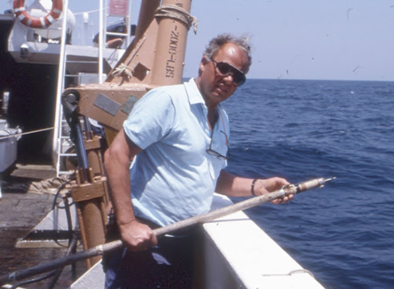 A scientist on the deck of a research ship at sea on a sunny day. He is holding a long pole with a sharp point at the end, used to insert tags into a shark’s fin. 