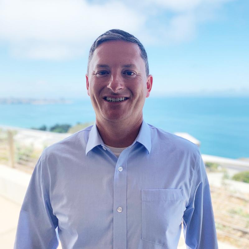 John Crofts wears a blue dress shirt and smiles at the camera with a sunny beach background.