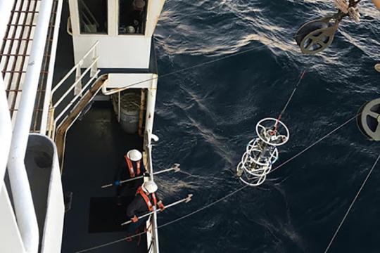 Aerial view of two crew members wearing hard hats at the rail on the main desk of the research vessel. They stand ready to snag a piece of water sampling equipment as it is raised from the water by a winch, using gaffs designed for this purpose.