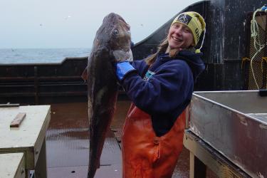 Photo of Ingrid Spies holding up a large Pacific cod on a boat deck.