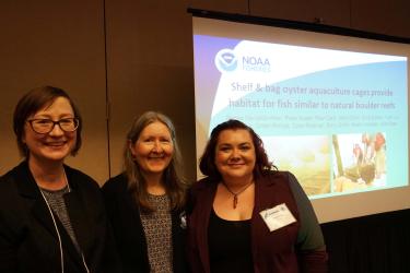 Three female scientists stand in front of a slide presentation in a conference room. The slide reads “Shelf & bag oyster aquaculture cages provide habitat for fish similar to natural boulder reefs”. 