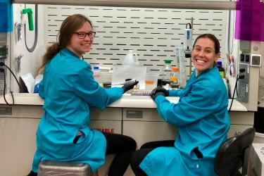 Two technicians seated in a lab wearing gloves and lab coats. They are turned to face the camera and smiling. 