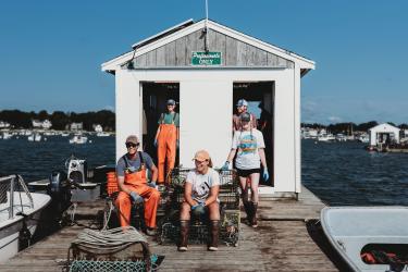 Five people in waders and baseball caps sit on oyster crates on a dock surrounded by blue water. 