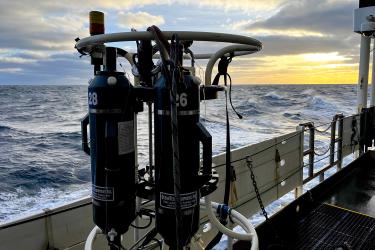 A large piece of ocean water sampling gear sits on the side deck of a research vessel while at sea. The ocean is choppy and the sunrise is peeking through the clouds.