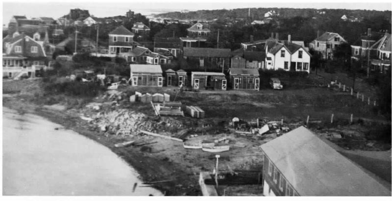 Three pigeon coops and sheds with screening are visible at the edge of a parking lot at center. View north shows private homes in the background looking toward Buzzards Bay. Great Harbor is at left. Materials and some small boats on the edge of the parking lot near the harbor at left. Photo: Courtesy Woods Hole Historical Museum Archives. 